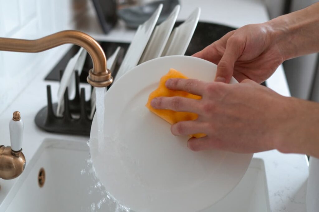 Imagem de uma pessoa usando luvas de borracha amarelas enquanto lava um prato com uma esponja verde, debaixo de uma torneira de cozinha. A água está correndo, e há espuma de sabão na esponja e no prato, ilustrando a tarefa de lavar louça como parte da rotina de limpeza doméstica. Esta imagem destaca a importância de manter a higiene ao lavar louça e pode servir de lembrete de áreas que muitas vezes são negligenciadas, como os cantos dos pratos e os utensílios de cozinha.